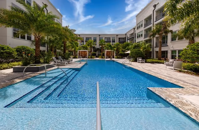Outdoor swimming pool surrounded by palm trees and lounge chairs, with a clear blue sky above and a multi-story building on both sides.