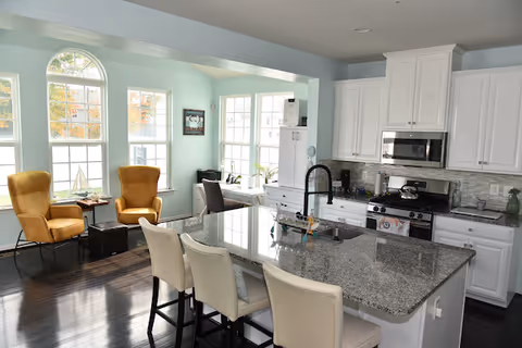 Bright kitchen area with a large granite island countertop featuring a sink and three white chairs. The kitchen has white cabinets, a stainless steel microwave, and a stove. In the background, there is a seating area with two mustard yellow armchairs, a small table, and large windows letting in natural light.