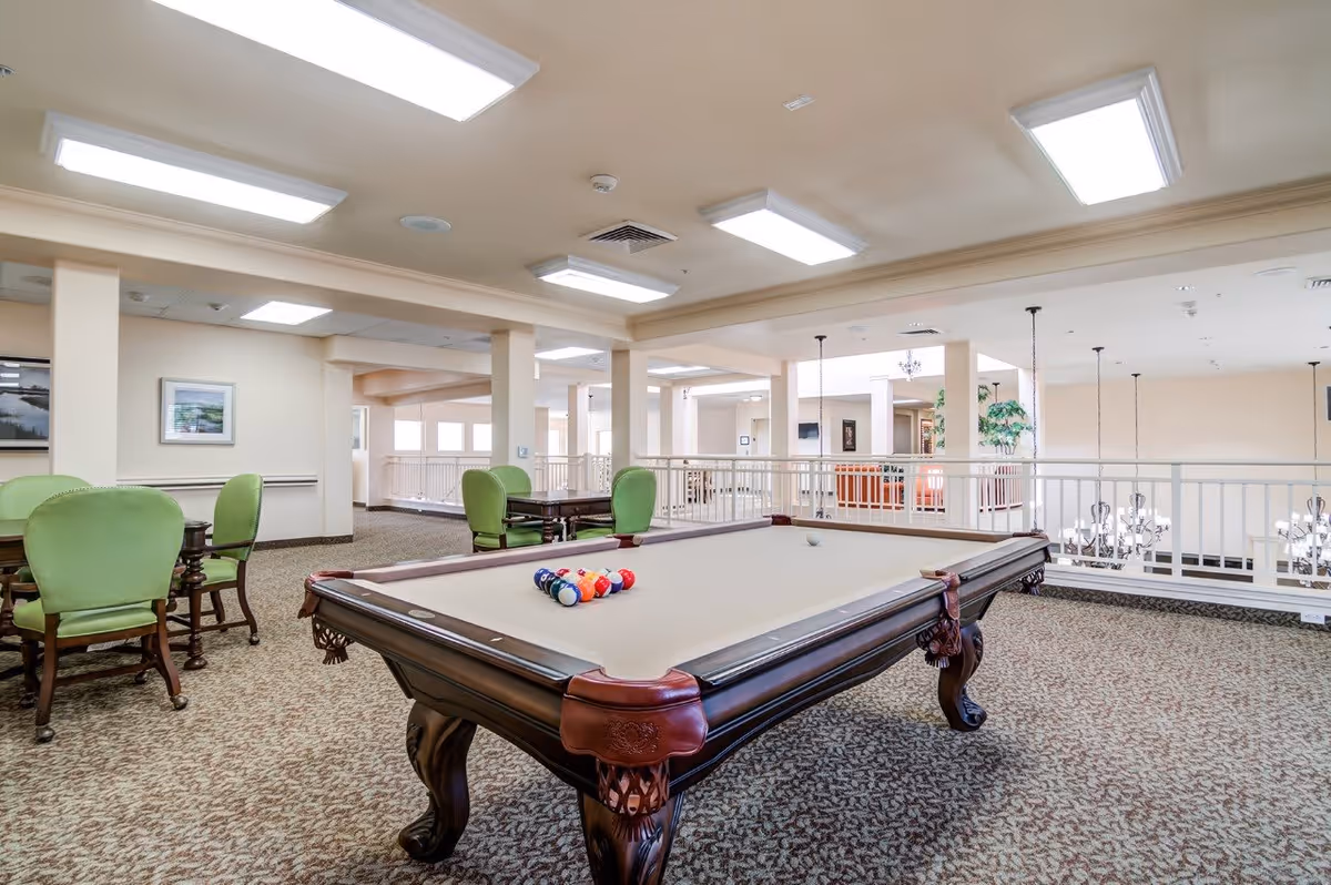 A spacious indoor recreational area featuring a billiards table with balls arranged for a game. Surrounding the table are several green cushioned chairs and small tables. The room has beige walls, carpeted floors, and bright overhead lighting. There is a railing overlooking a lower level with chandeliers visible below.