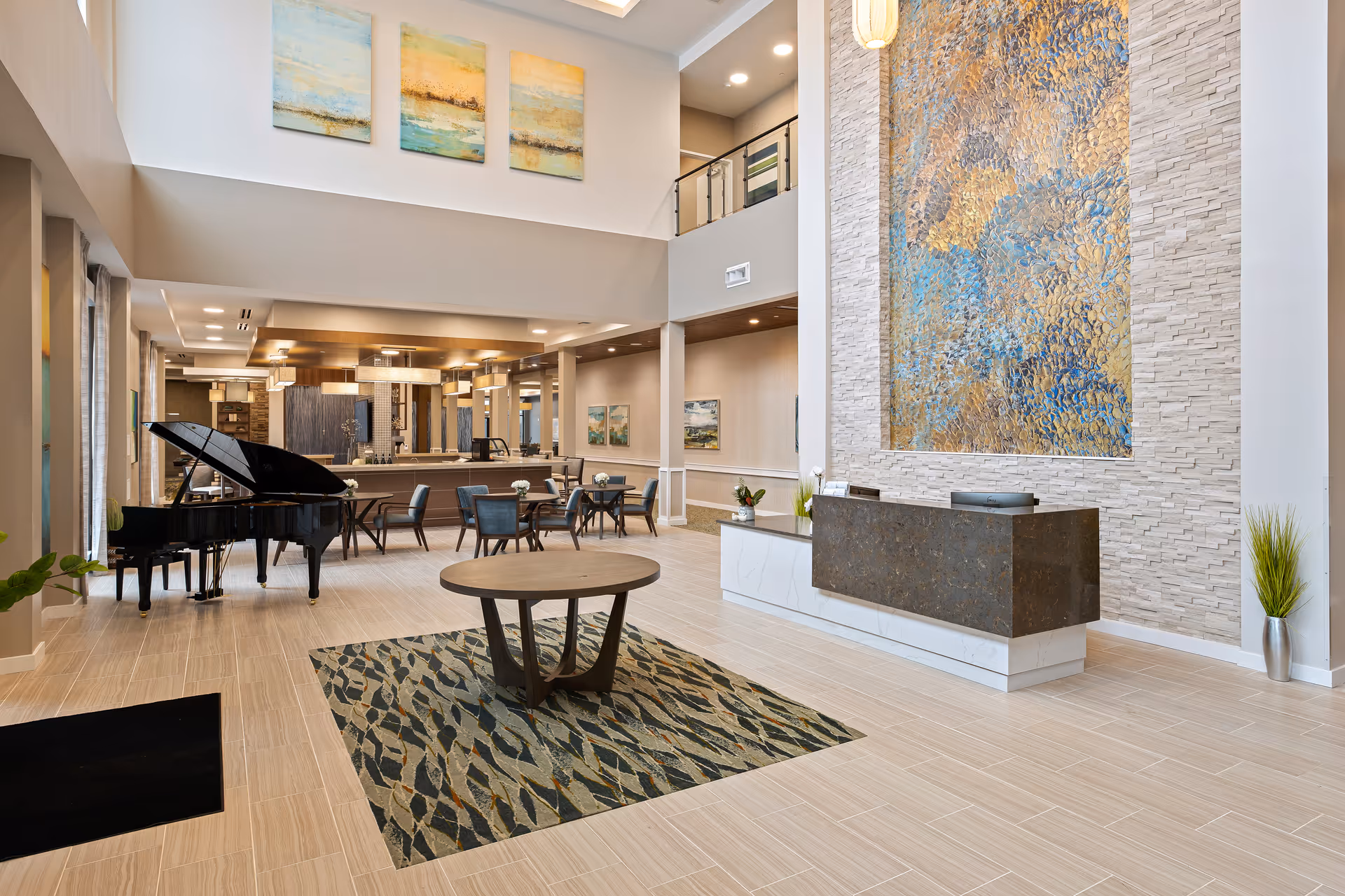 Spacious and modern senior living facility common area with a grand piano on the left, several small round tables with chairs, a large decorative wall art piece behind a reception desk, and a high ceiling with three abstract paintings on the upper wall.