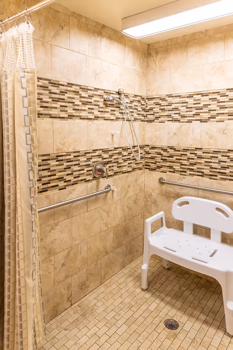 Accessible tiled shower with grab bars, a white shower chair, handheld showerhead, and a shower curtain.