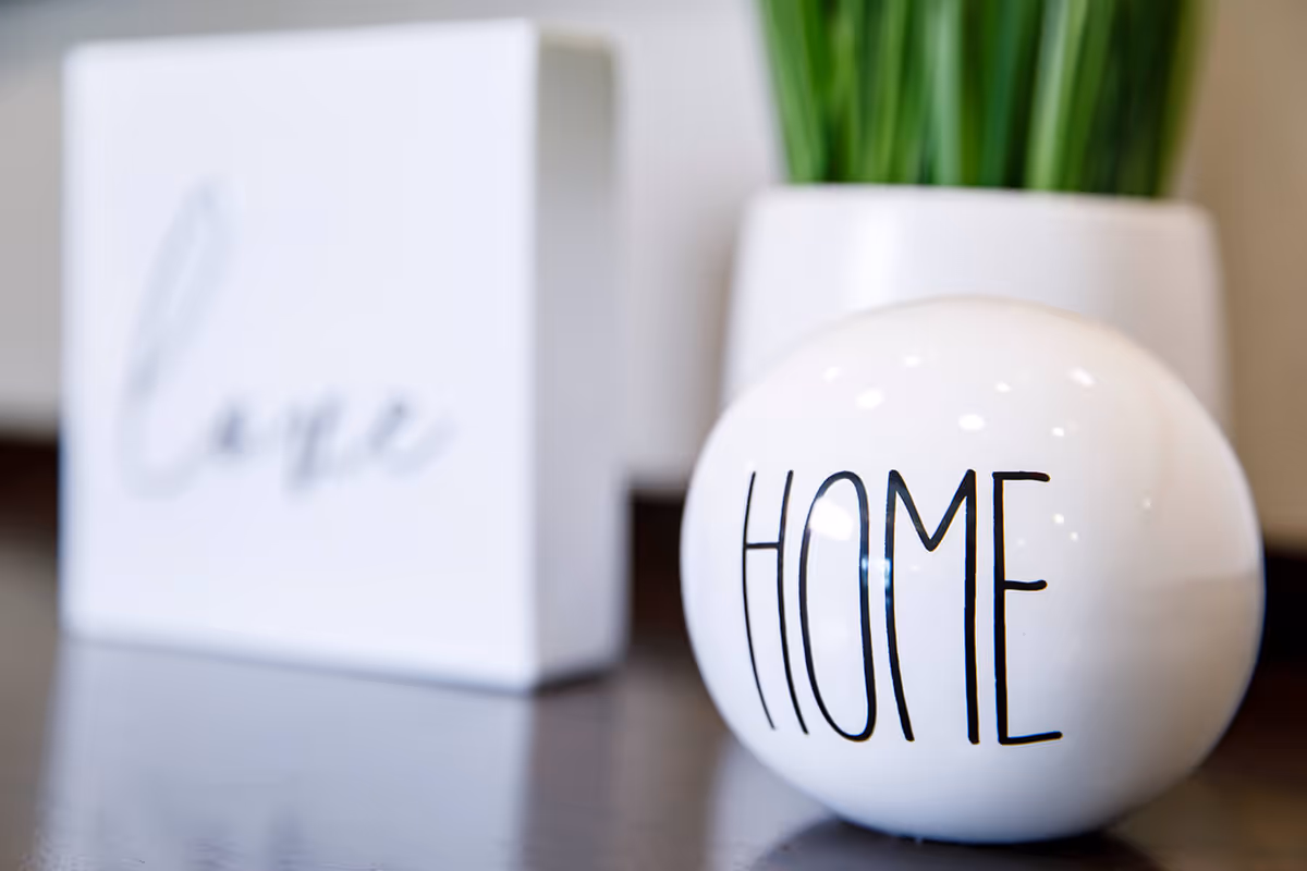 Close-up of a decorative white sphere labeled "HOME" on a tabletop with a blurred "love" sign and a potted plant in the background.