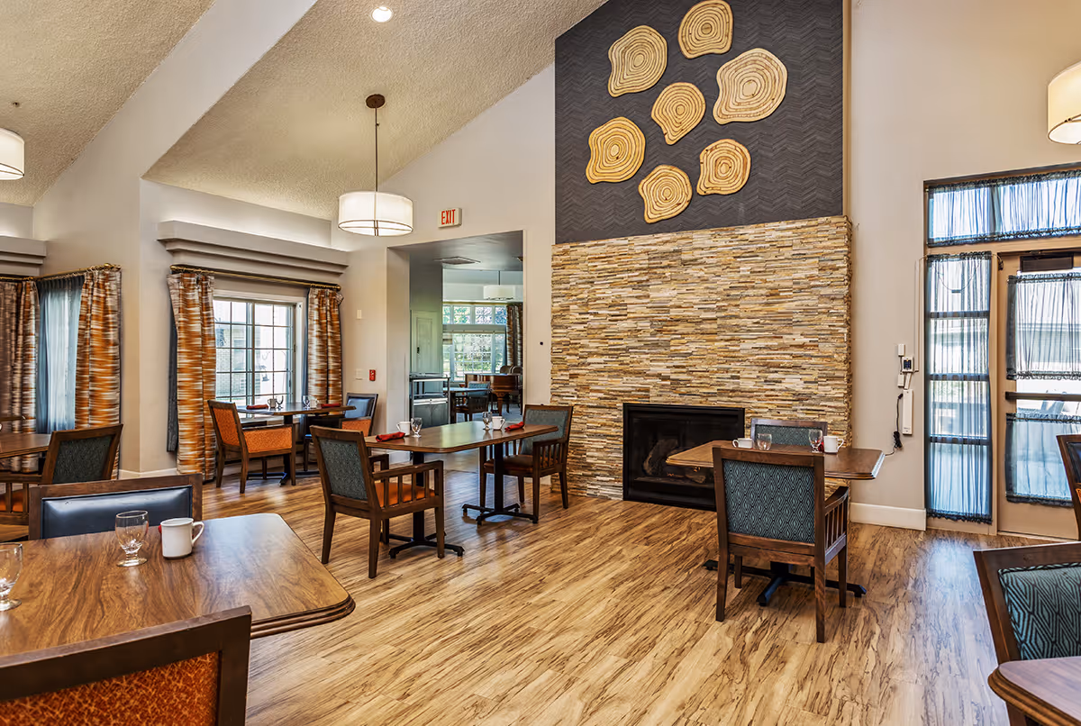 A dining room with wooden tables and chairs, set with glasses and cups. The room features a stone fireplace with decorative wooden art above it, large windows with patterned curtains, and a door with sheer curtains. The floor is wooden and the ceiling has modern light fixtures.