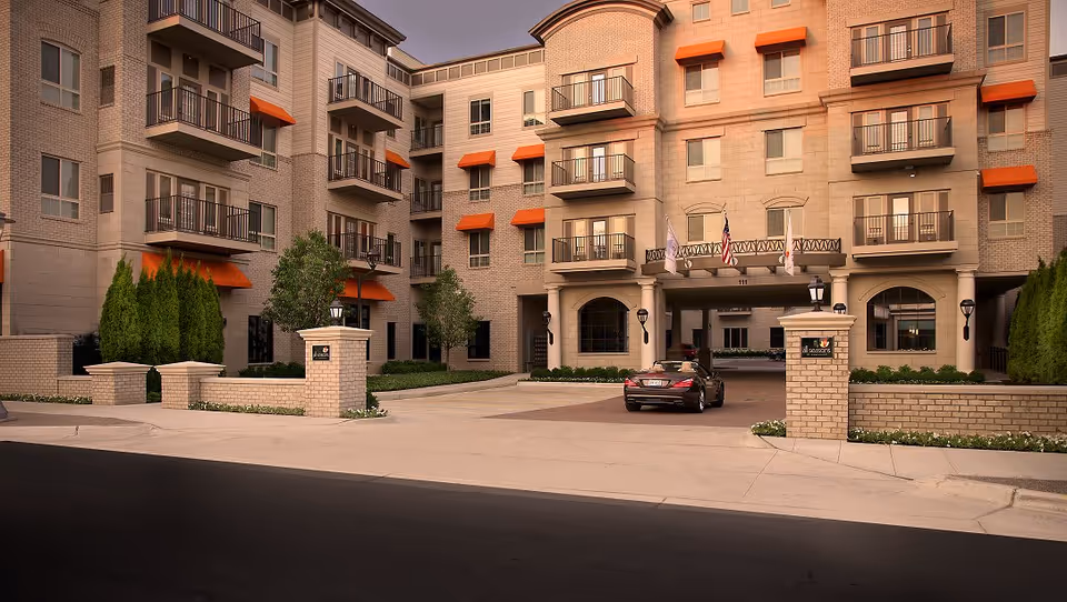 Exterior view of a multi-story senior living facility named All Seasons Birmingham, featuring beige brick walls, multiple balconies with orange awnings, a driveway entrance with brick pillars, and a black car parked near the entrance under a covered area.