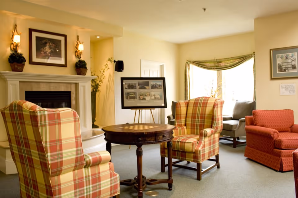 A cozy living room area with two plaid armchairs facing a round wooden table. There is a fireplace with a framed picture and two wall sconces above it. In the background, there is a window with a green valance, a gray sofa, and a red armchair. A framed collage of photos is displayed on an easel near the doorway.