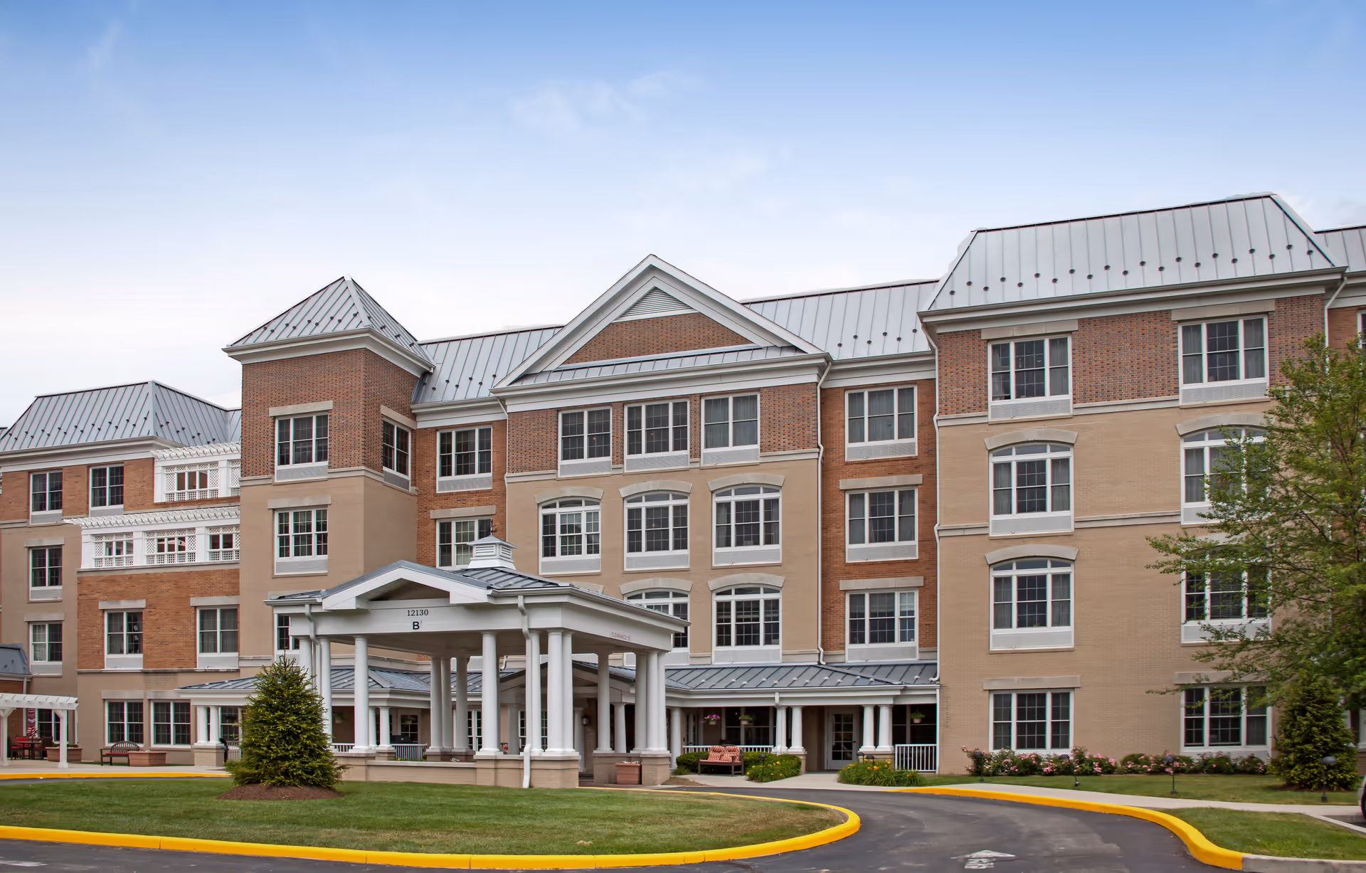 Front exterior of a multi-story brick and stucco senior living building with a covered porte-cochère and circular driveway.