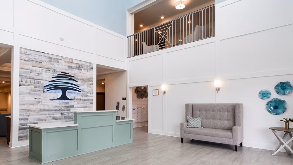 A bright and spacious reception area with a light green front desk, a gray tufted loveseat with a patterned pillow, and decorative blue wall art. The walls are white with paneling, and there is a large logo of a stylized tree on a wood-textured accent wall behind the desk. Above, there is a balcony railing overlooking the space.