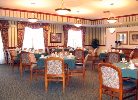 A dining room with multiple round tables covered with green tablecloths, each set with cups, glasses, and silverware. The room has patterned wallpaper, floral curtains on the windows, framed artwork on the walls, and wooden chairs with patterned upholstery. Ceiling lights hang above the tables, and there is a sideboard with coffee dispensers and cups in the background.