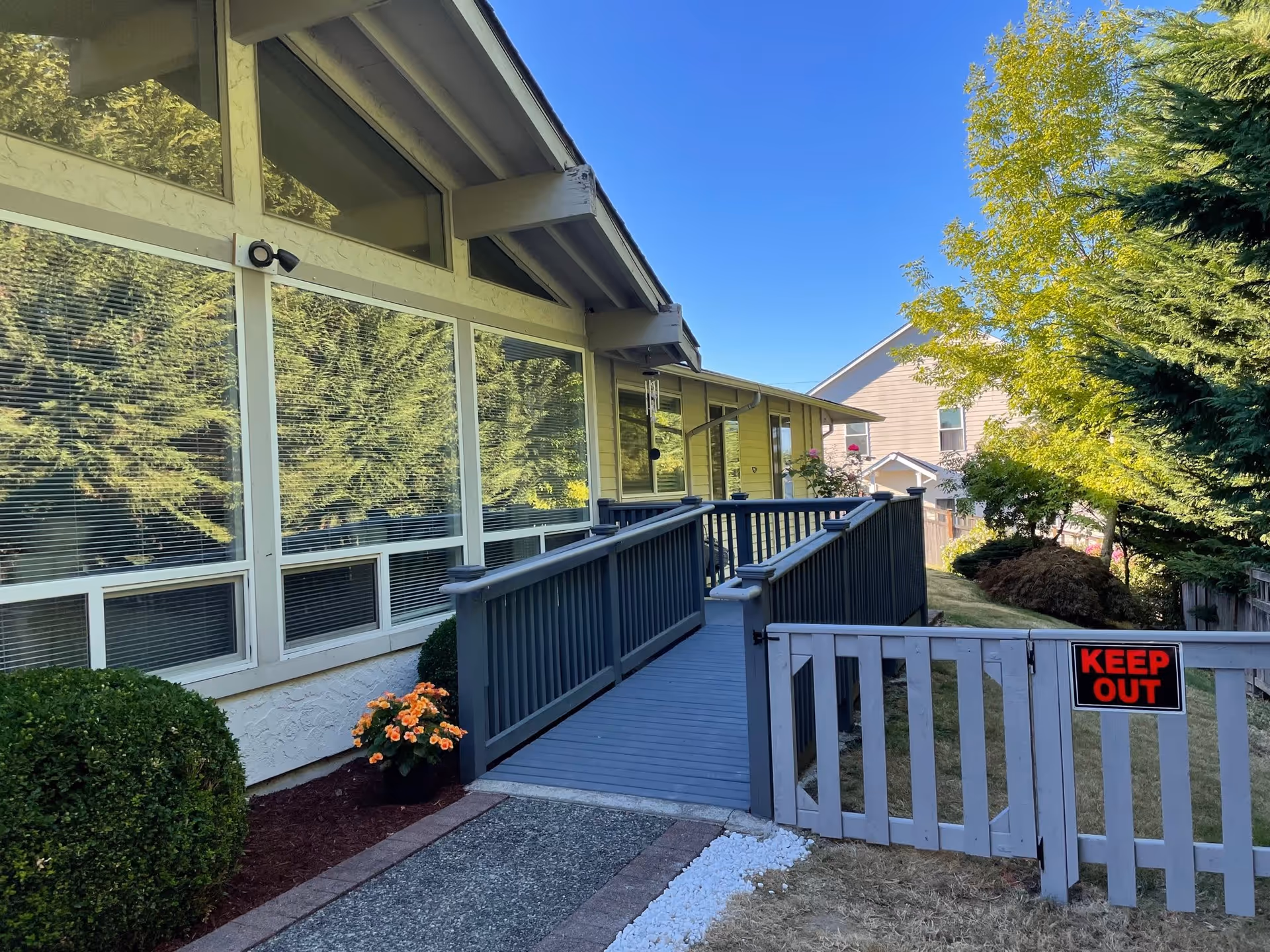 Exterior view of a single-story building with large windows and a gray wooden ramp leading to the entrance. There is a small garden with bushes and flowers on the left side and a wooden gate with a red and black 'KEEP OUT' sign on the right. Trees and another house are visible in the background under a clear blue sky.