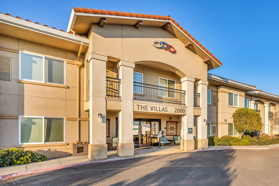 Exterior view of a two-story senior living facility building named The Villas with the address number 2000. The building has beige walls, a red-tiled roof, multiple windows, and a covered entrance with seating. There are some bushes and a tree near the entrance under a clear blue sky.