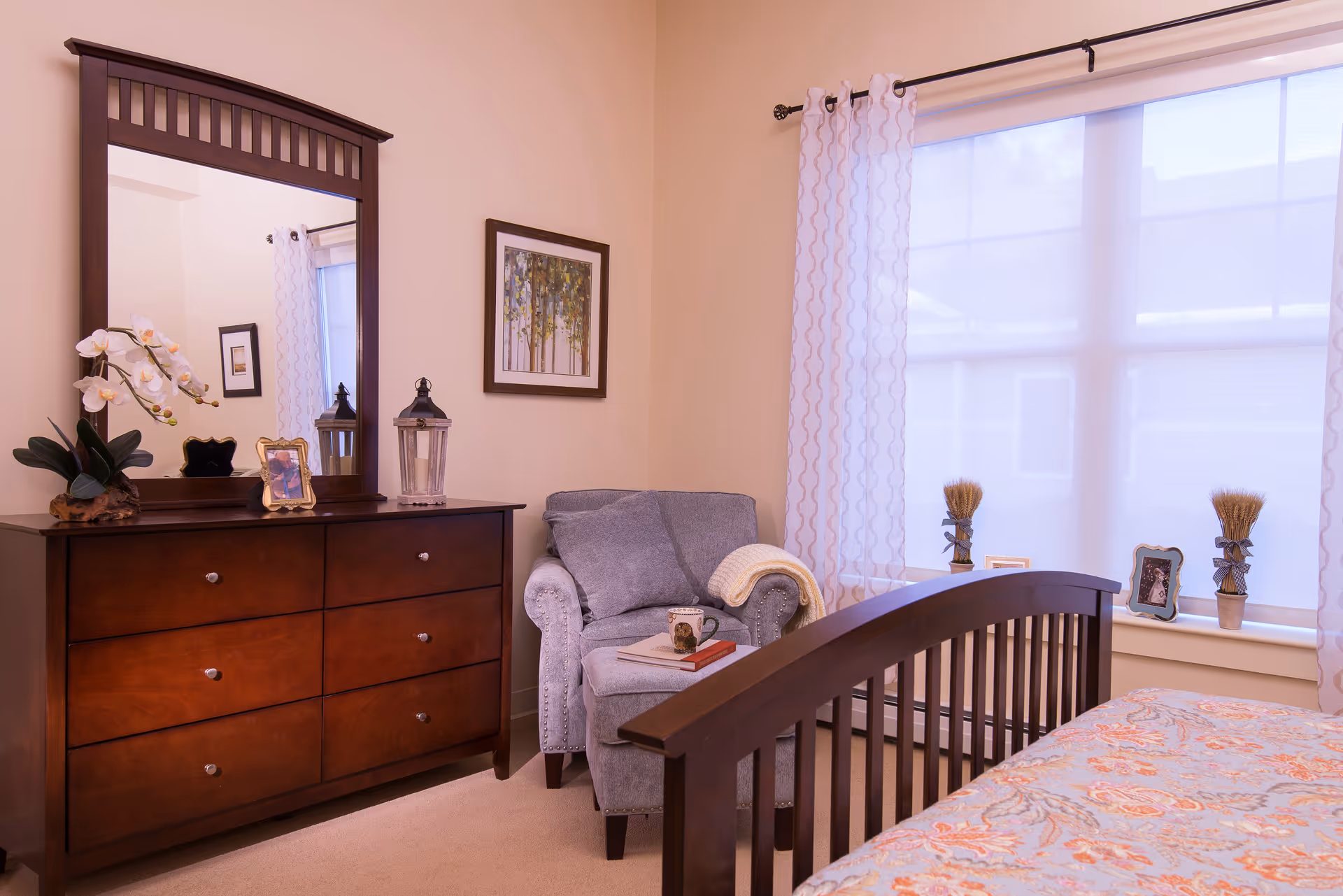 A cozy bedroom featuring a wooden dresser with a large mirror, a framed picture on the wall, a comfortable gray armchair with a blanket and a book on it, and a bed with a patterned bedspread. The room has a large window with sheer curtains allowing natural light to enter.