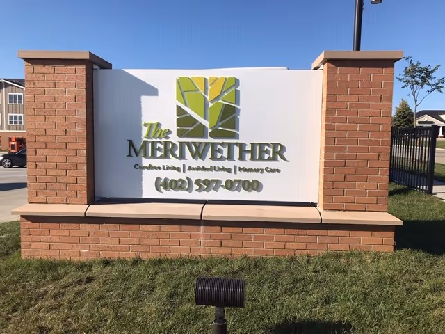 A brick and white sign for The Merriwether senior living facility, displaying services including Carefree Living, Assisted Living, and Memory Care, along with a phone number (402) 597-0700. The sign is outdoors on a grassy area with a clear blue sky in the background.