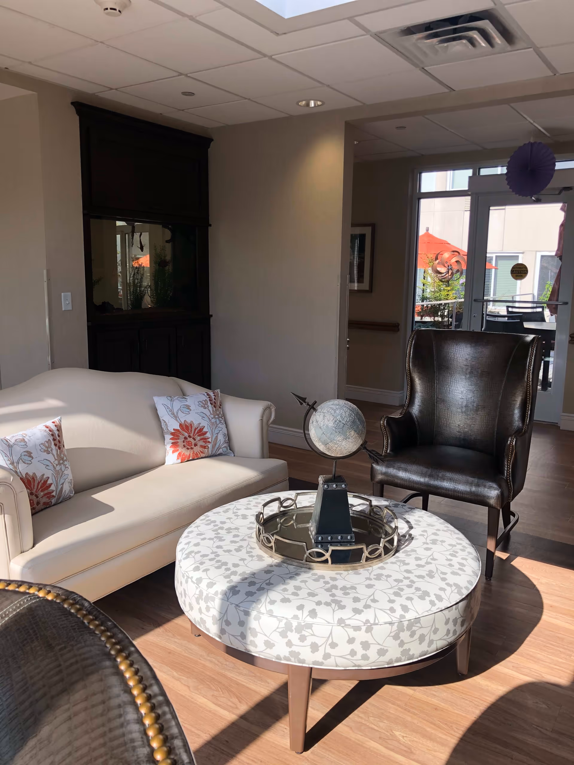 A cozy senior living facility lounge area with a cream-colored sofa adorned with floral pillows, a dark leather armchair, and a round ottoman with a floral pattern. On the ottoman is a decorative globe on a stand. The room has wooden flooring and a large windowed door leading to an outdoor patio with red umbrellas.
