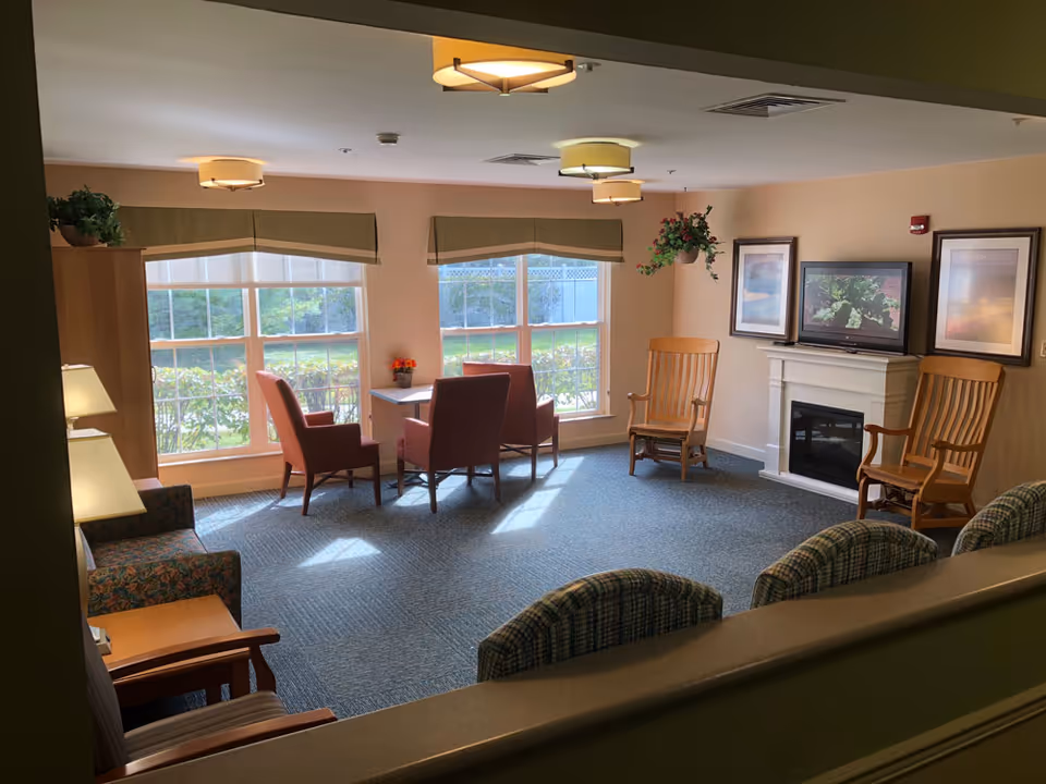 A cozy living room area with large windows letting in natural light. The room features a small table with four chairs near the windows, two wooden rocking chairs near a white fireplace with a TV mounted above it, and additional seating including a patterned armchair and three cushioned chairs in the foreground. The walls are painted a soft beige color, and there are framed pictures and a hanging plant adding to the decor.