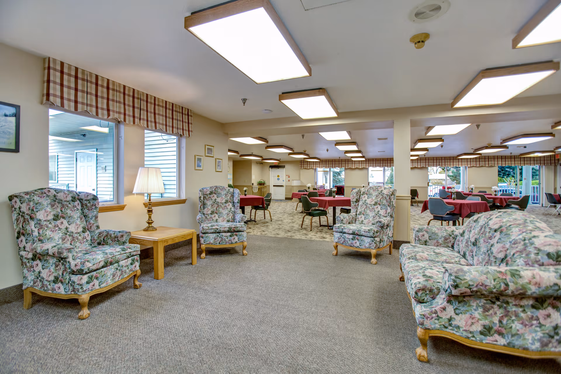 A senior living facility common area with floral upholstered armchairs and a sofa arranged around a wooden side table with a lamp. In the background, there is a dining area with tables covered in red tablecloths and chairs, illuminated by ceiling lights. Windows with plaid valances allow natural light into the space.