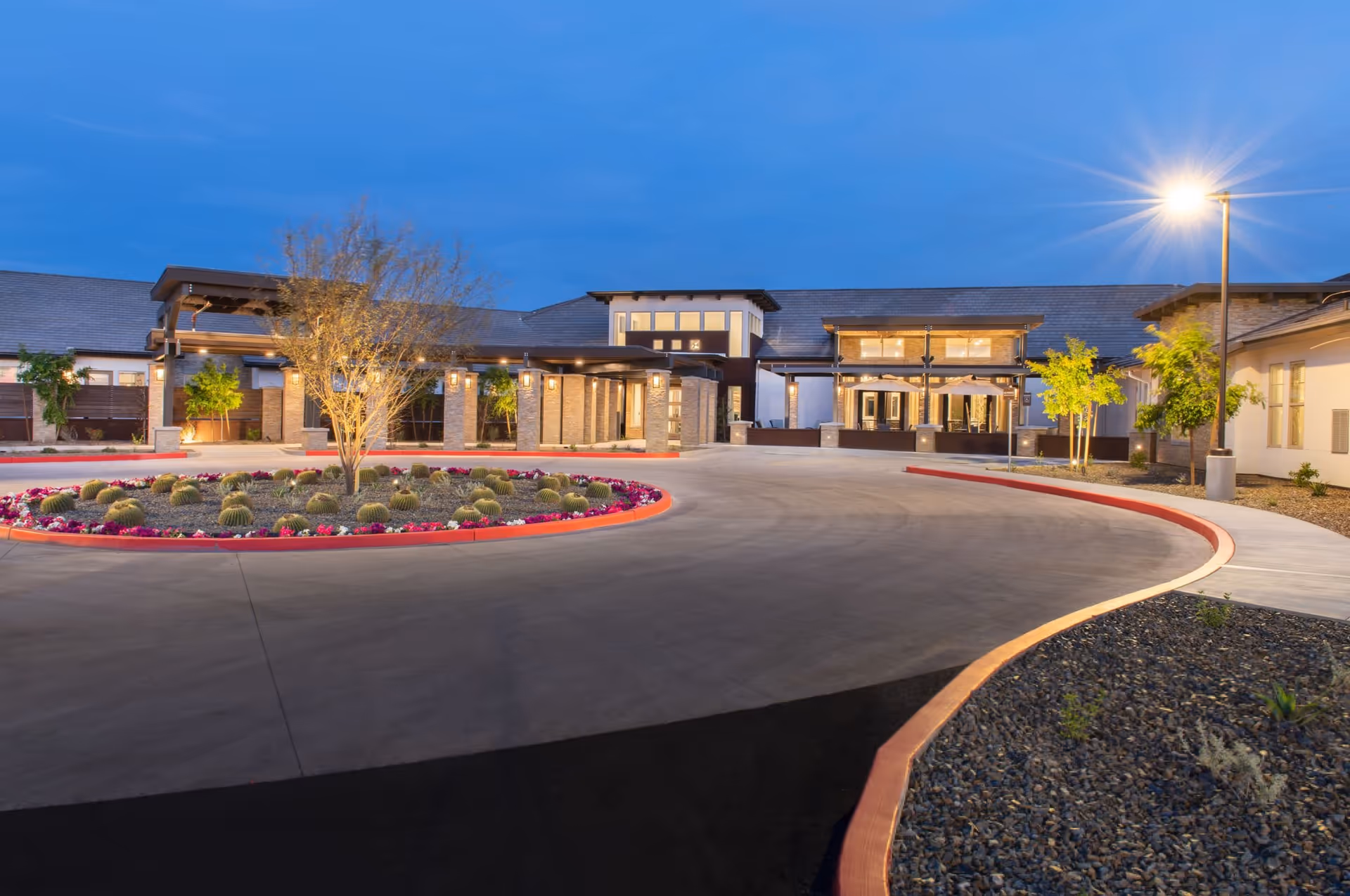 Exterior view of Spring Gardens Senior Living Peoria at dusk, showing a well-lit entrance with a circular driveway, landscaped with cacti and flowers, and a modern building with large windows and a covered walkway.