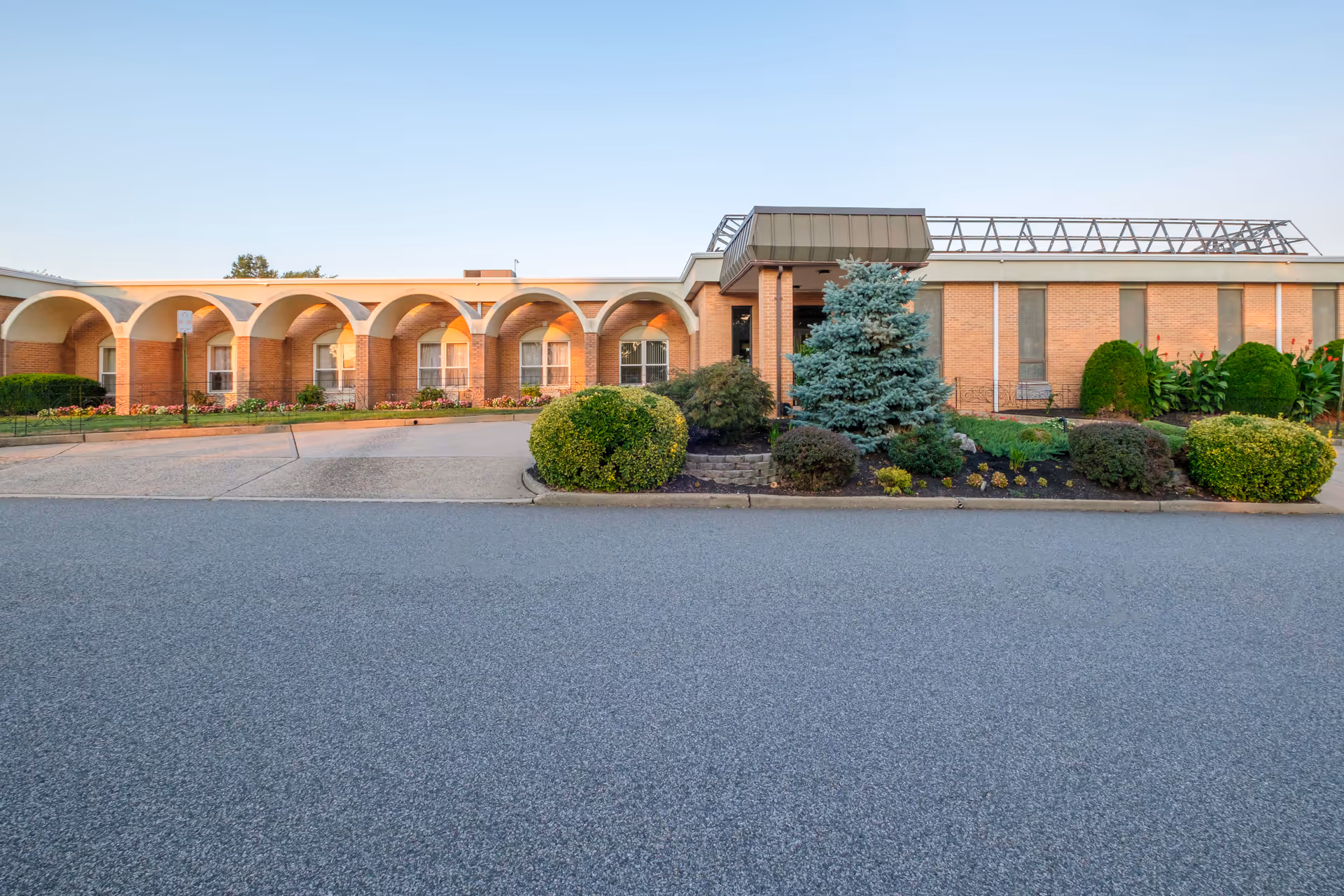Exterior front view of St Joseph's Senior Home - Assisted Living and Nursing Center, showing a single-story brick building with arched architectural features, windows, and landscaped bushes and trees in front under a clear sky.