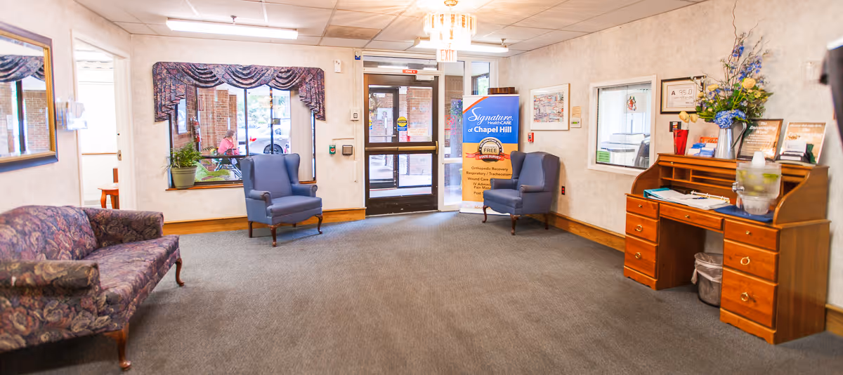 A waiting or lobby area in a senior living facility with a floral patterned couch on the left, two blue armchairs near the entrance door, a wooden desk with flowers and documents on the right, and a large window with a valance. A banner near the door reads 'Signature HealthCARE of Chapel Hill.'