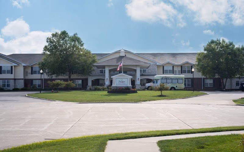 Front exterior view of Bickford of Bloomington facility with a driveway, green lawn, trees, and a sign in front. The building is two stories with a covered entrance and an American flag flying above the sign.