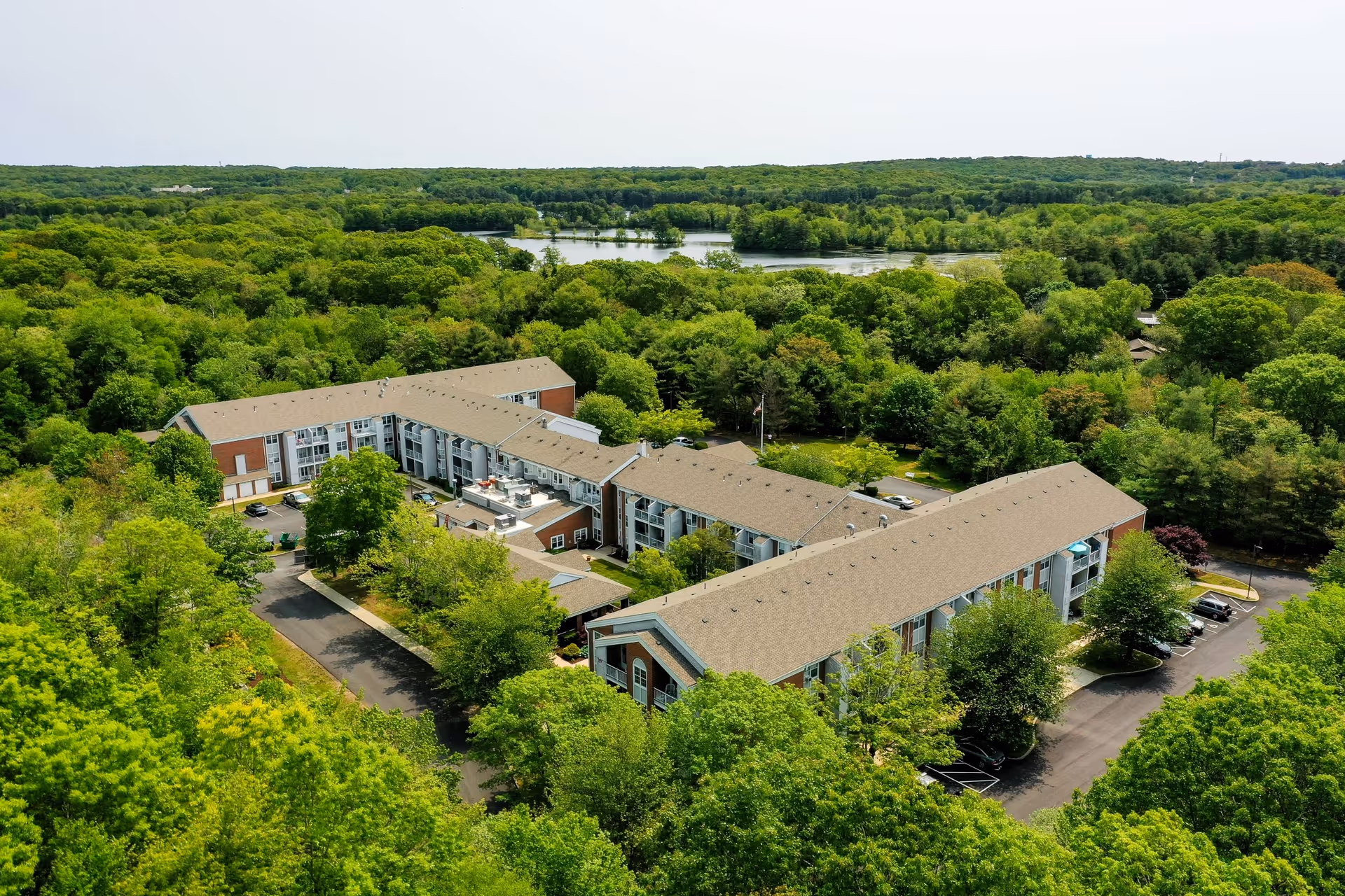 Aerial view of Solstice Senior Living at Groton, a large L-shaped building surrounded by dense green trees with a lake visible in the background under a clear sky.