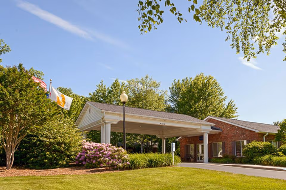 Exterior view of Life Care Center of Hendersonville showing a brick building with a covered entrance, surrounded by green trees, bushes, and flowering plants under a clear blue sky.
