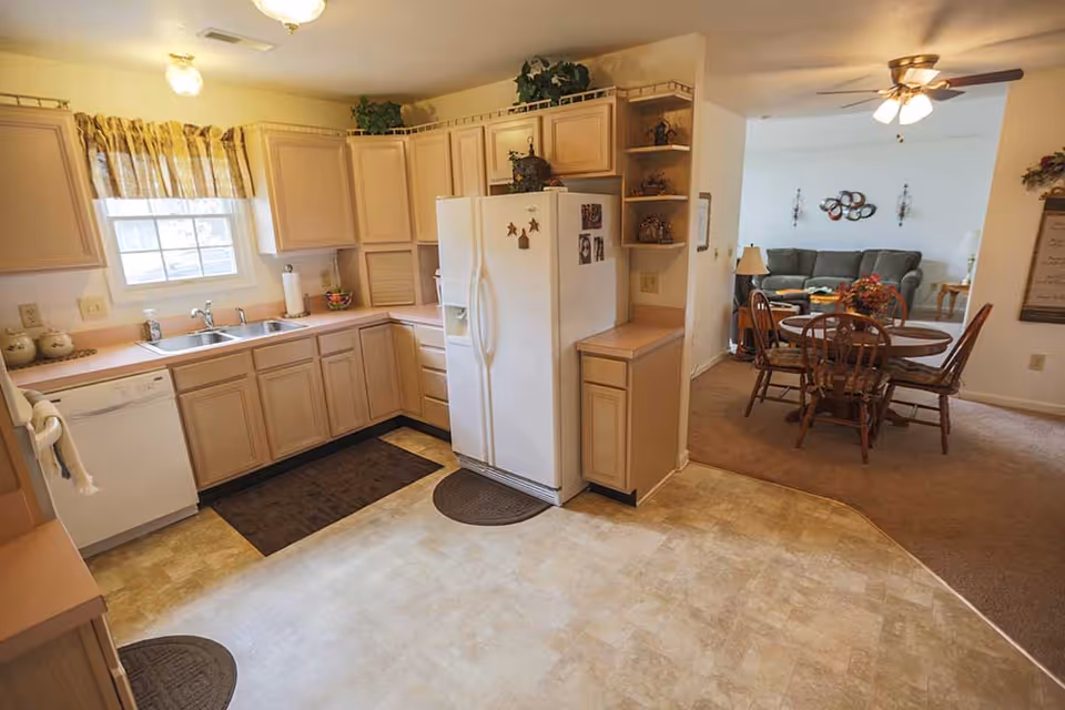 A bright kitchen with light wood cabinets, a white refrigerator, dishwasher, and double sink under a window with yellow curtains. The kitchen opens into a dining area with a wooden round table and four chairs, and a living room with a gray couch and wall decorations. The floor in the kitchen is tiled, while the dining and living areas are carpeted.