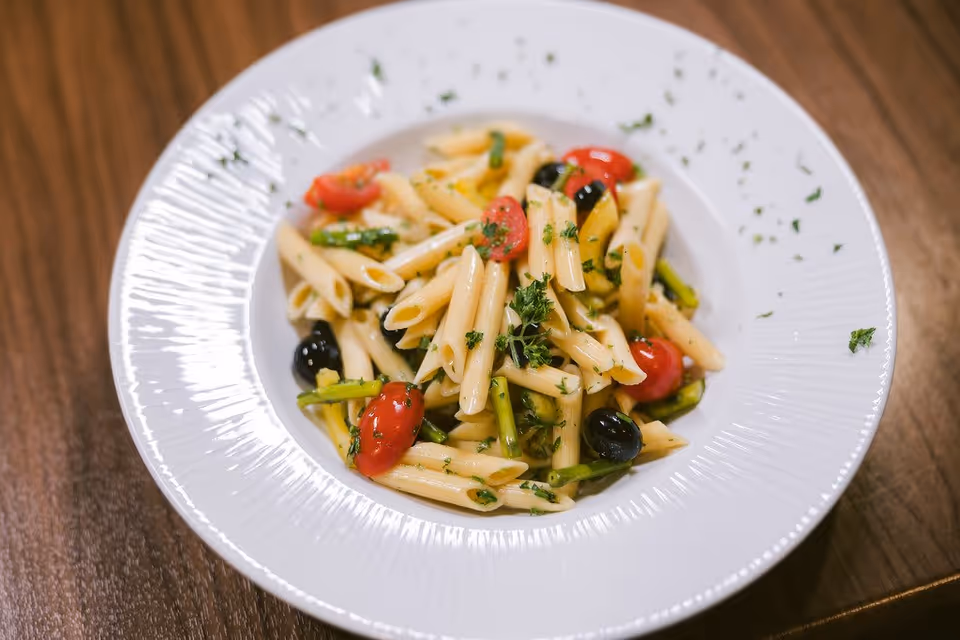 A white plate filled with penne pasta mixed with cherry tomatoes, black olives, asparagus, and garnished with chopped parsley, placed on a wooden table.