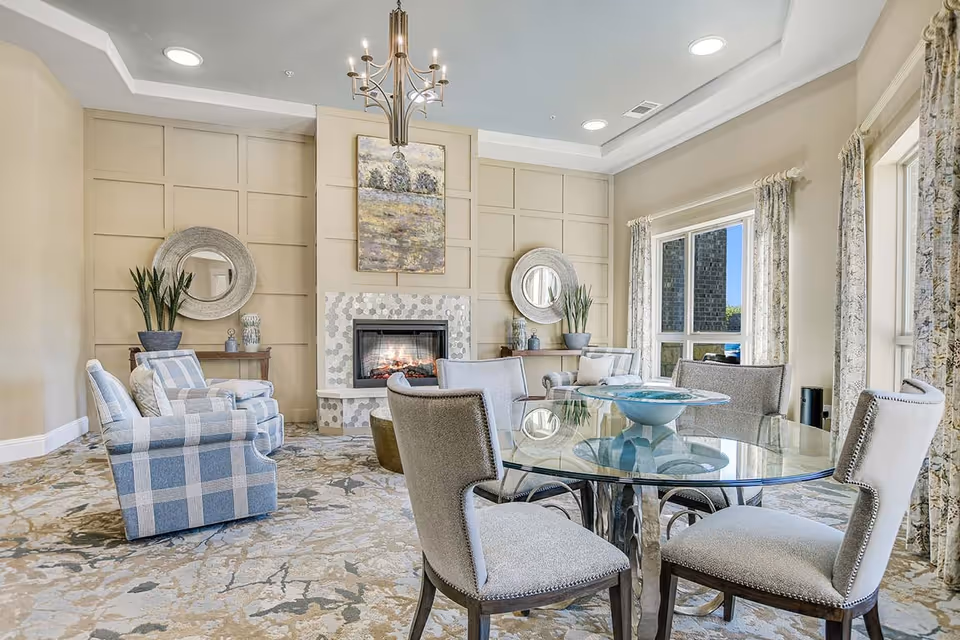 Sunlit communal living room with a fireplace, plaid armchairs, a round glass dining table and decorative mirrors.