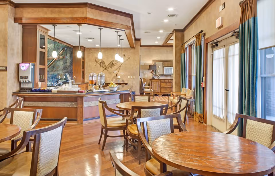 A dining area in a senior living facility with round wooden tables and cushioned chairs. The room features a serving counter with beverage dispensers and a water cooler. Pendant lights hang from the ceiling, and large windows with curtains allow natural light to enter. The decor includes warm tones and wooden accents.