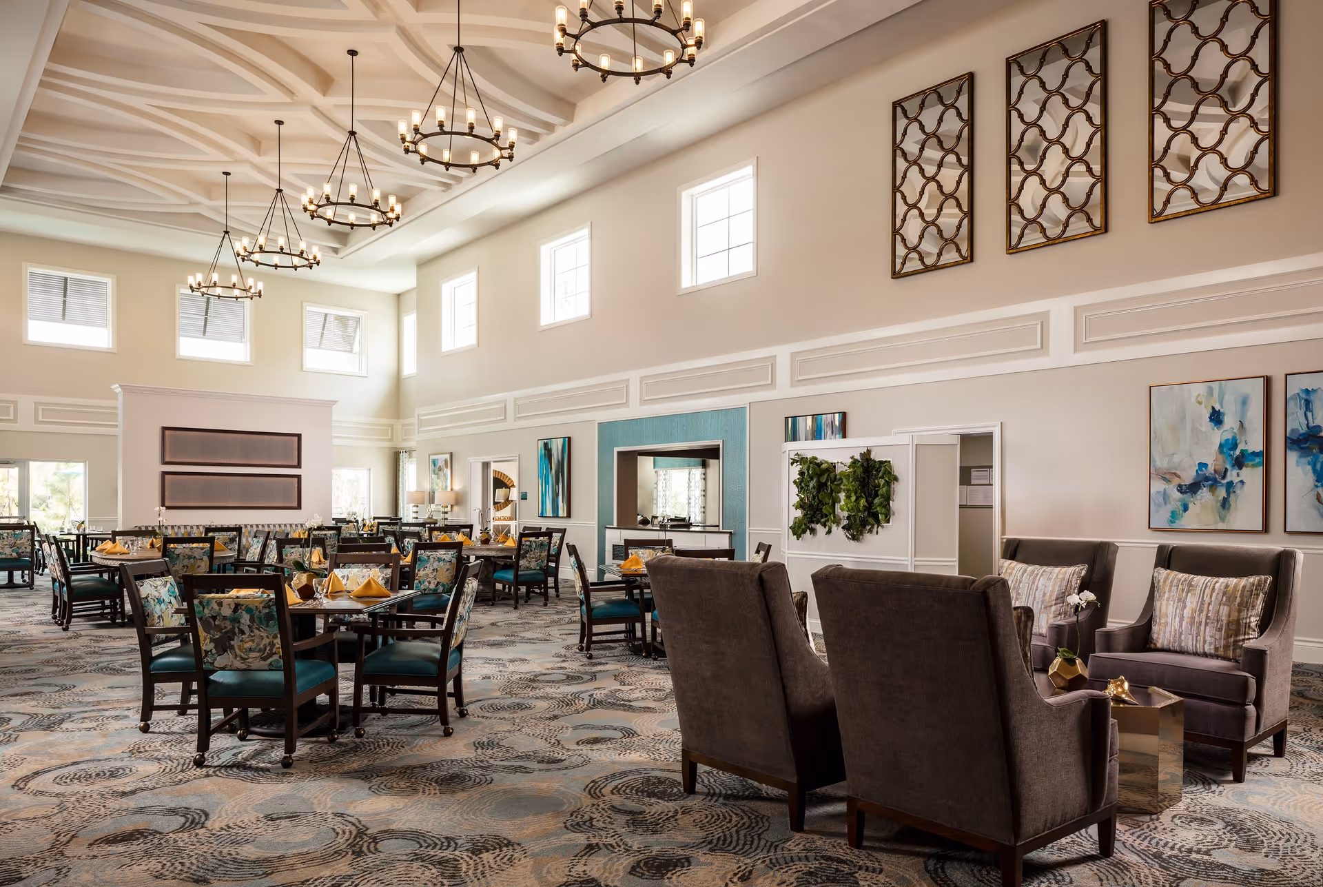 Spacious dining room with multiple tables and chairs, featuring floral upholstery and teal cushions. The room has high ceilings with decorative beams and hanging chandeliers. There are large windows letting in natural light, abstract artwork on the walls, and a seating area with gray armchairs and a small table.