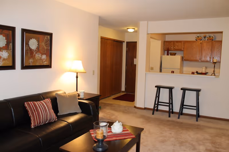 Cozy living room with a black leather sofa, coffee table and lamp, opening to a kitchen counter with two bar stools.