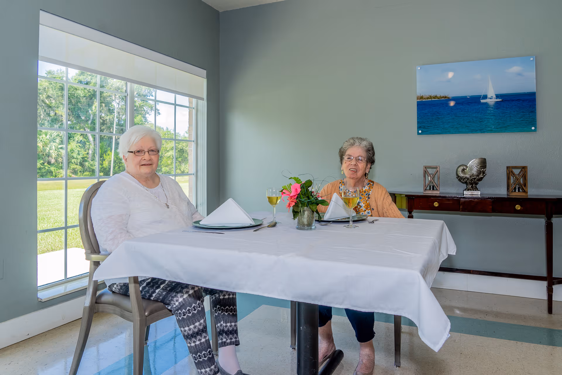Two elderly women sitting at a dining table with a white tablecloth, set with plates, napkins, and glasses of a yellow beverage. They are in a room with a large window showing green outdoor scenery and a wall with a framed picture of a sailboat on the ocean. A wooden sideboard with decorative items is visible behind them.