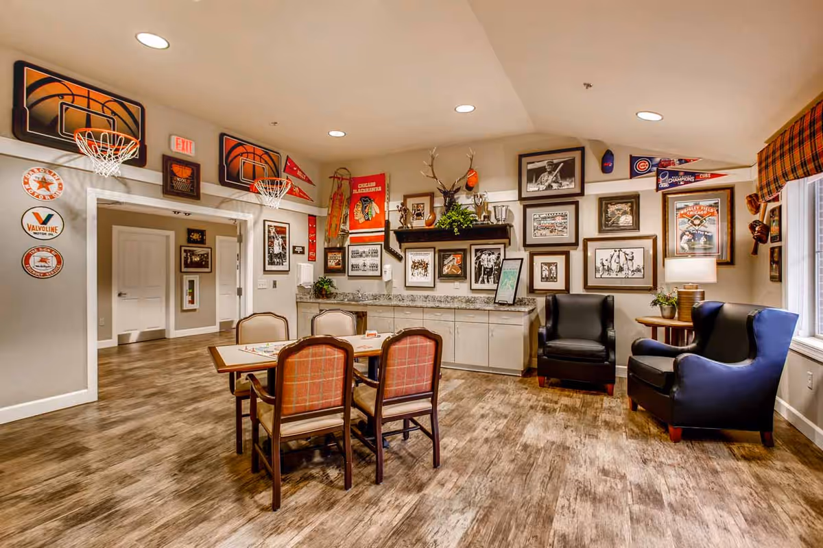 A cozy common area in a senior living facility decorated with sports memorabilia including basketball hoops, framed photos, and pennants. The room features a table with four chairs in the center, two black armchairs near a window with plaid curtains, and a countertop with cabinets along the back wall. The floor is wood-style, and the walls are painted beige.