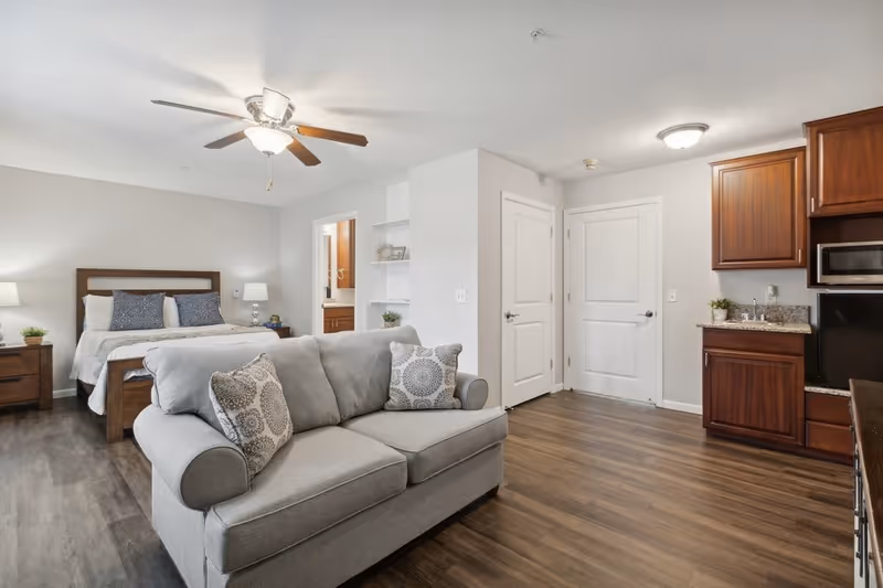 A spacious studio apartment with a gray sofa in the foreground, a bed with pillows and nightstands with lamps in the background, a ceiling fan with light, wooden flooring, and a kitchenette with wooden cabinets and a microwave on the right side.
