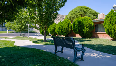 A peaceful outdoor area at Skyline Ridge Nursing and Rehabilitation Center featuring a black metal bench on a concrete pathway surrounded by green grass, trees, and neatly trimmed bushes with a brick building in the background under a clear blue sky.