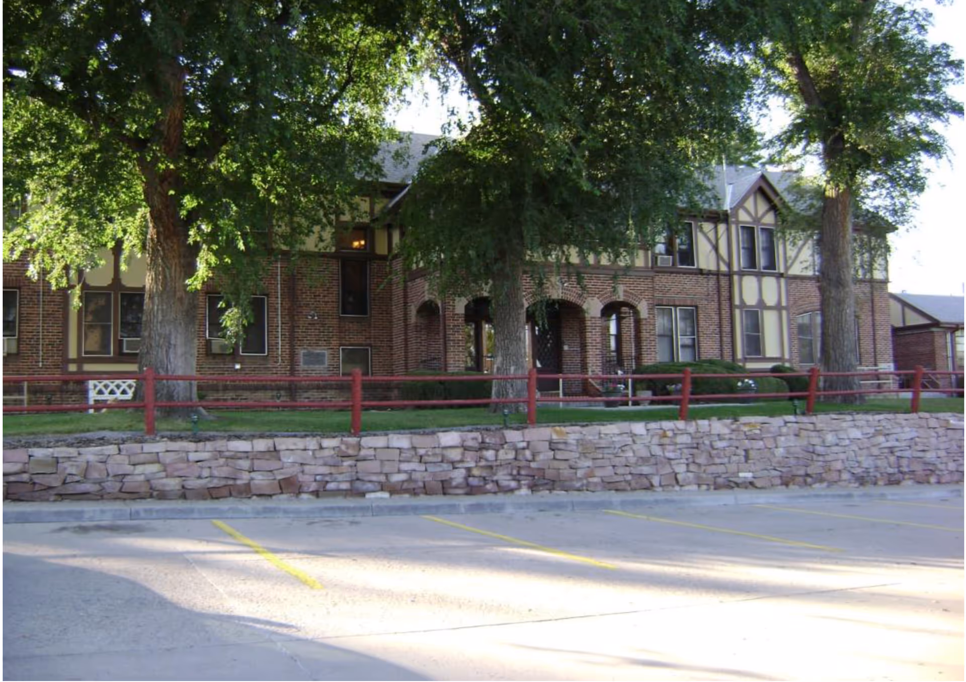 Brick Tudor-style two-story building with large trees, a red fence and stone retaining wall in front of a parking area.