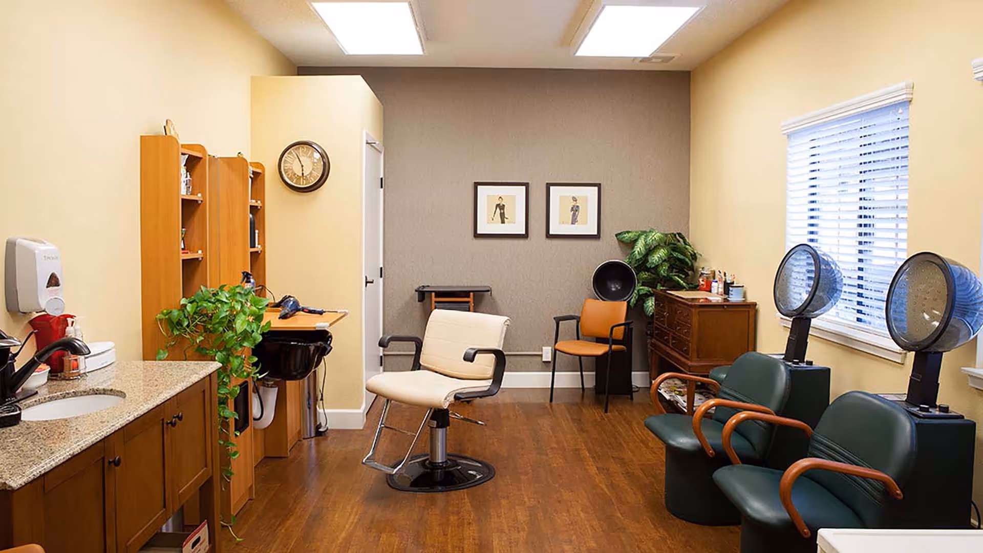 Interior of a hair salon room with wooden flooring, a beige salon chair in the center, two green chairs with hair dryers on the right, a wooden cabinet with hair products, a plant, a wall clock, and framed pictures on the back wall.