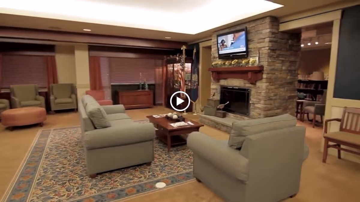 A cozy living room area with two gray sofas facing a stone fireplace with a mounted TV above it. There is a wooden coffee table on a patterned rug between the sofas. In the background, there are two armchairs, a round ottoman, and a wooden cabinet with window blinds behind it. The room has warm lighting and a welcoming atmosphere.