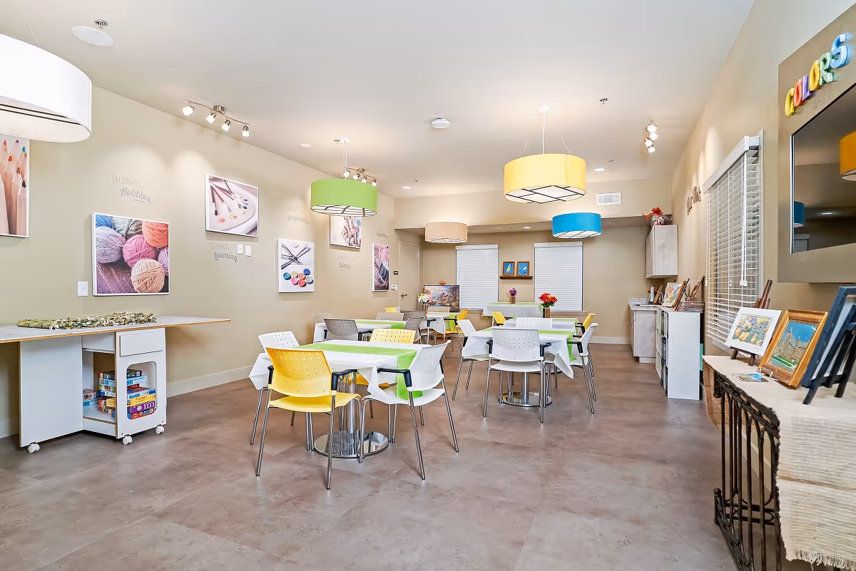 A bright and colorful activity room in a senior living facility with tables and chairs arranged for group activities. The walls are decorated with pictures of yarn, paintbrushes, and buttons, emphasizing hobbies and creativity. There are colorful hanging lamps in green, yellow, beige, and blue. A table on the left holds board games, and a side table on the right displays framed artwork. The room has a clean, welcoming atmosphere.