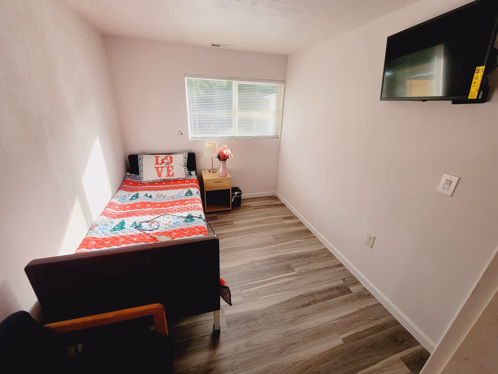 A small bedroom with a single bed covered in a red and white patterned blanket and a pillow with the word LOVE on it. Next to the bed is a wooden nightstand with a lamp and a vase of flowers. A window with blinds is above the nightstand. A flat-screen TV is mounted on the wall opposite the bed. The floor has wood-like vinyl planks.
