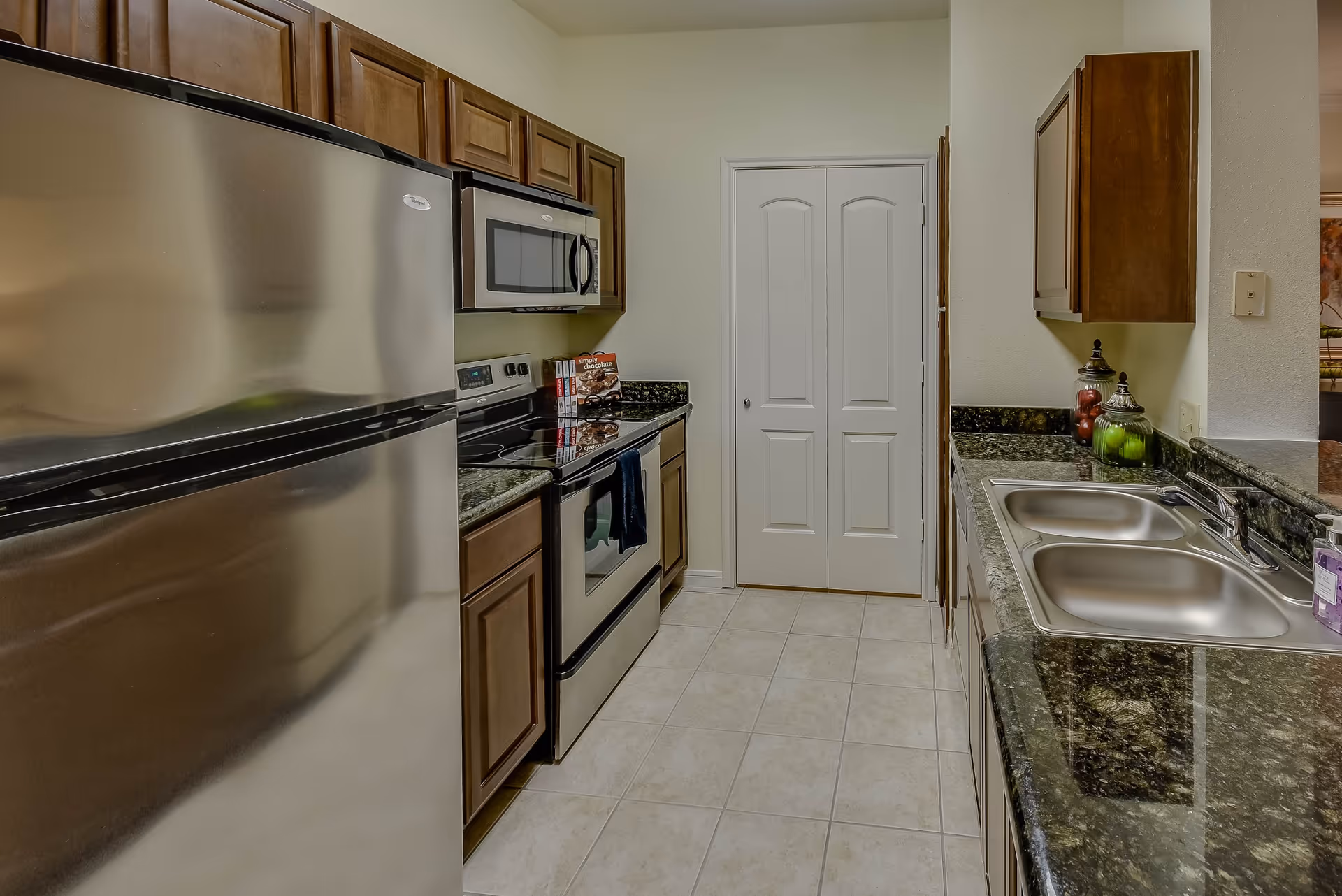 A modern kitchen with stainless steel appliances including a refrigerator, microwave, and electric stove. The kitchen has dark granite countertops, wooden cabinets, a double sink, and a tiled floor. There are decorative jars with fruit on the counter and a closed white double door at the end of the kitchen.