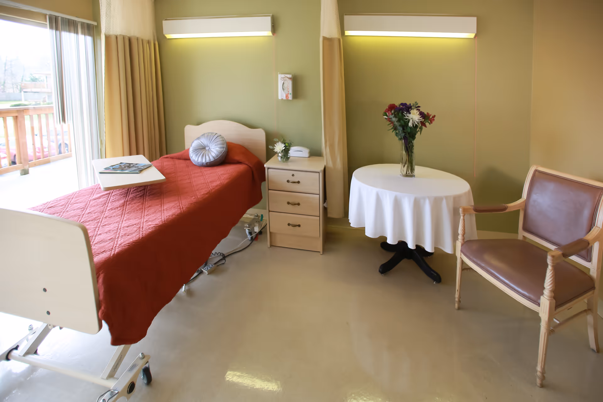 A senior living facility bedroom with a hospital-style bed covered with a red quilt and a round decorative pillow. Next to the bed is a small wooden nightstand with a telephone and a small vase with flowers. A round table with a white tablecloth and a vase of colorful flowers is beside a wooden chair with brown cushions. The room has light green walls and a large window with curtains letting in natural light.