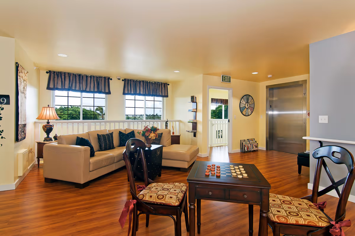 A cozy senior living common area with a beige sectional sofa adorned with dark pillows, a wooden coffee table with a checkers game set, two wooden chairs with patterned cushions, a table lamp on a side table, large windows with blue valances letting in natural light, and an elevator door in the background.