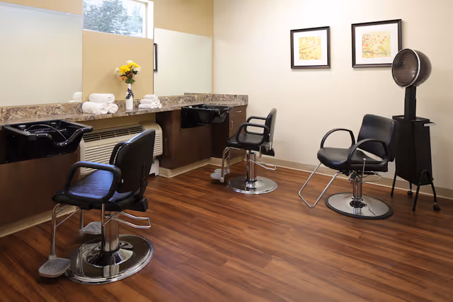 Interior of a salon room with three black salon chairs, two black hair washing sinks, a hair dryer, a countertop with a vase of yellow flowers and rolled white towels, and two framed pictures on the wall.
