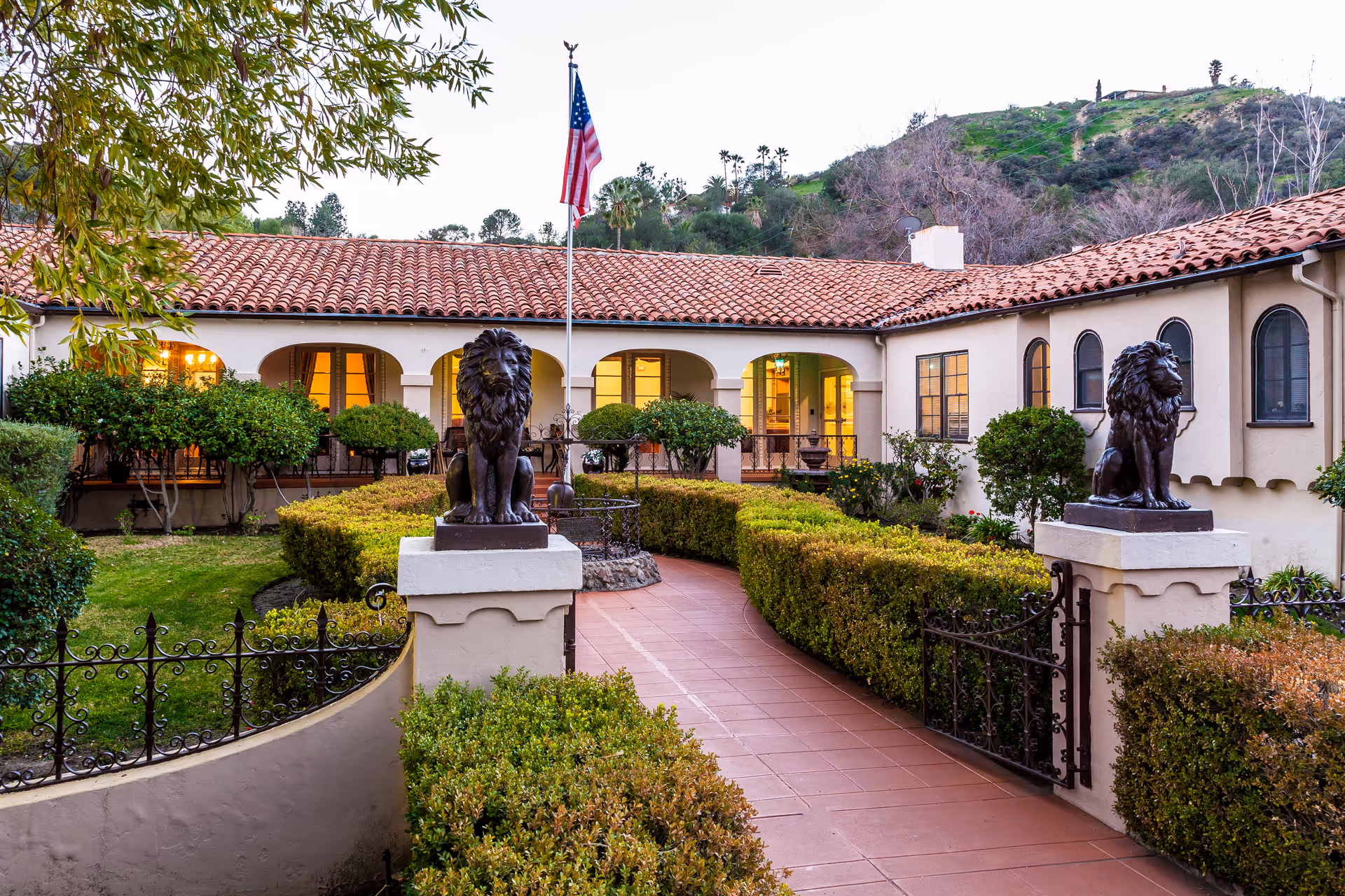 Entrance pathway to a retirement facility with two lion statues on pedestals flanking the walkway, manicured hedges and bushes on either side, a flagpole with an American flag in the center, and a building with arched windows and a red tile roof in the background during early evening.
