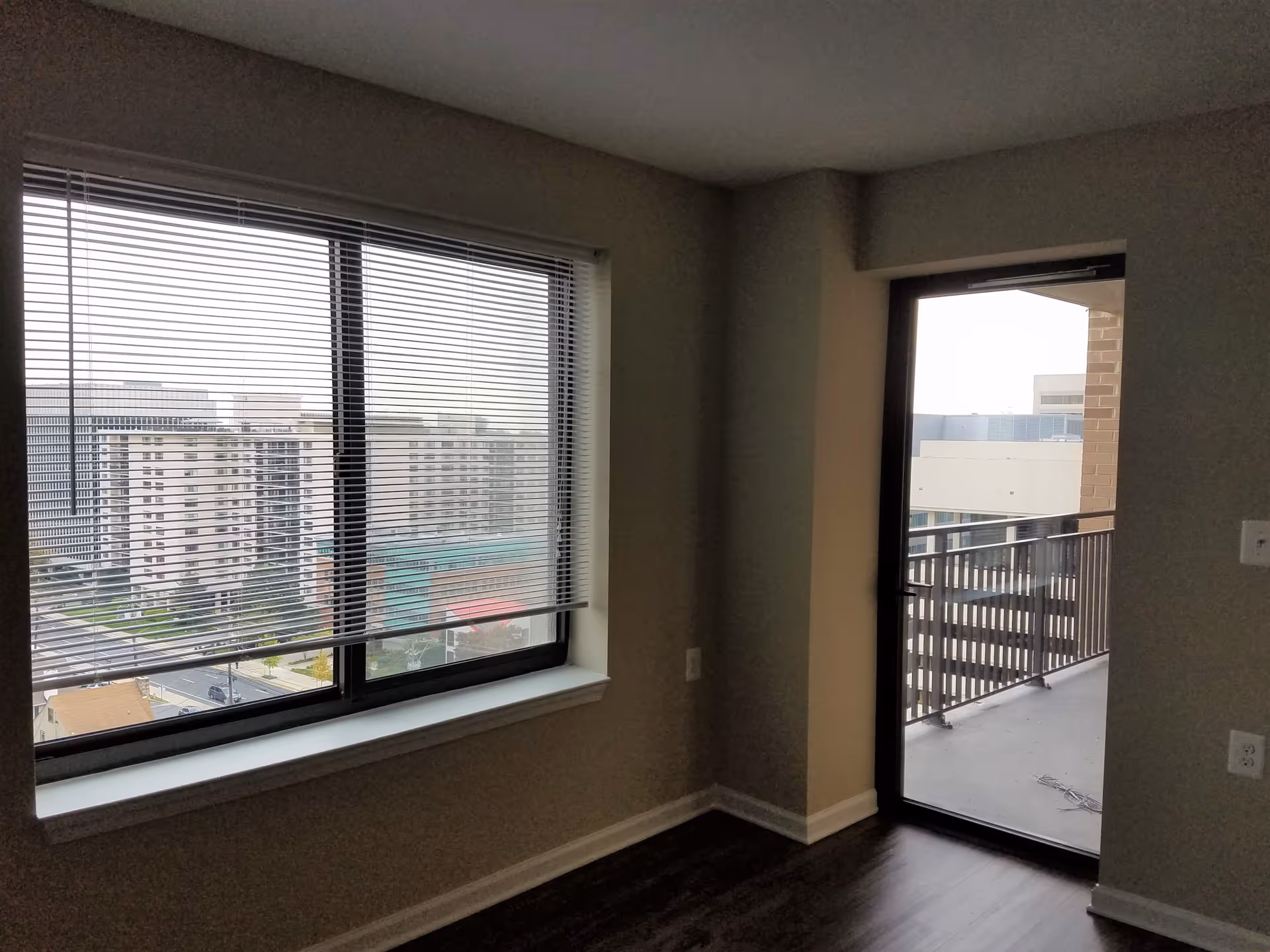 Interior corner of an apartment with a large window covered by horizontal blinds and a glass door leading to a balcony. Outside, several tall buildings are visible under an overcast sky.