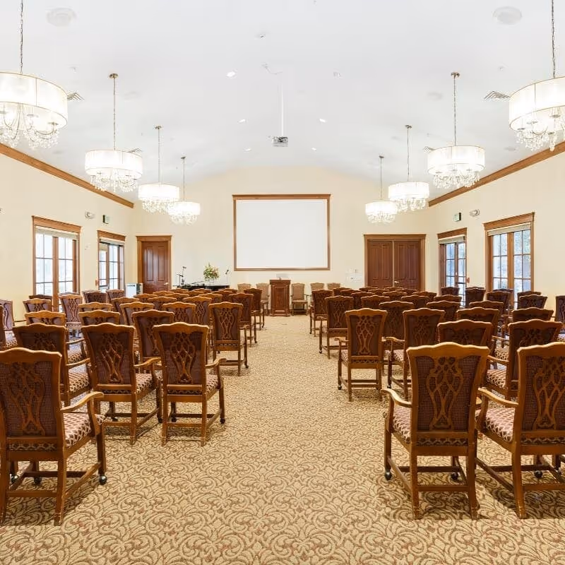 A large, well-lit meeting room with rows of ornate wooden chairs facing a projector screen and podium.