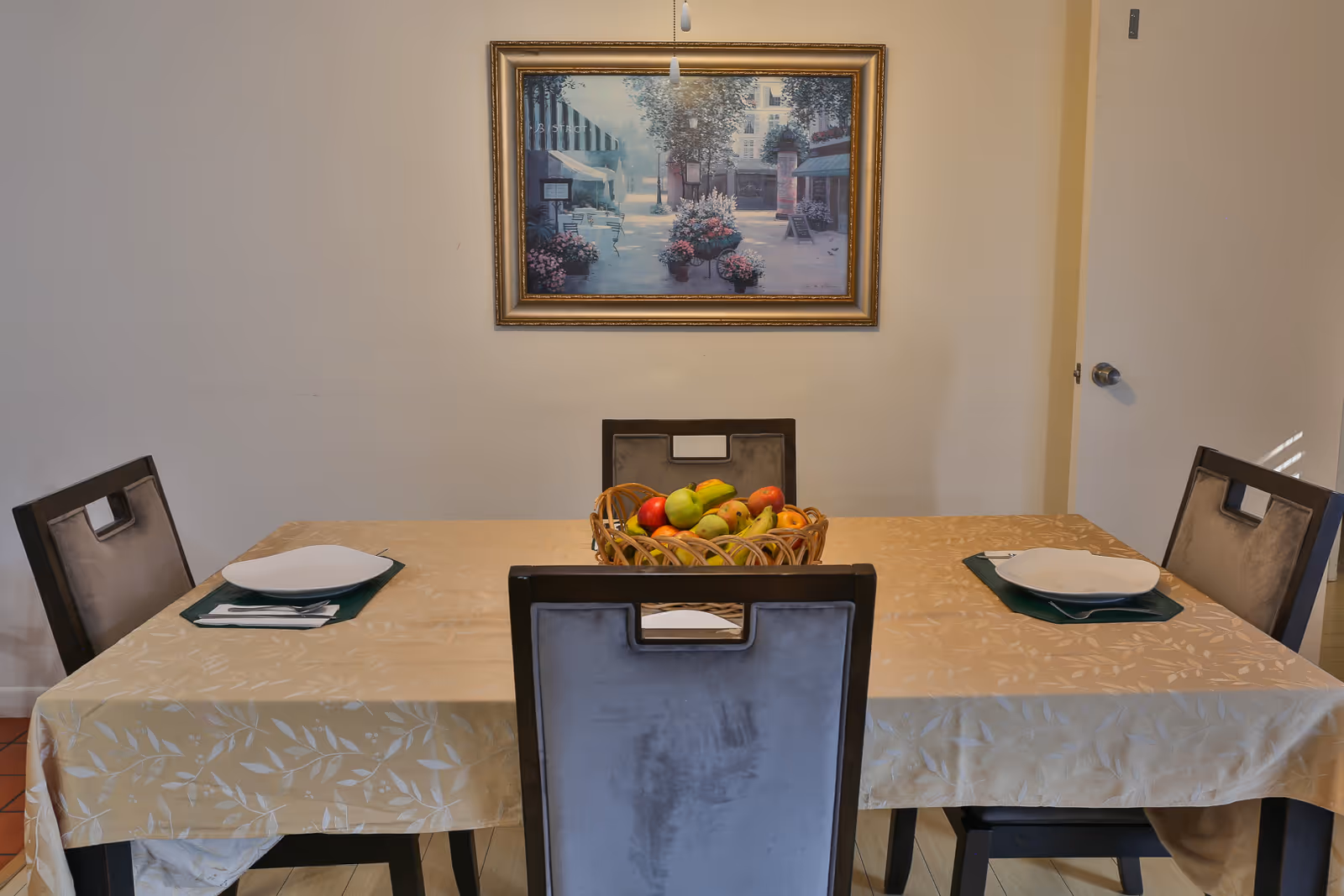 A dining table set for two with a beige tablecloth, two place settings with white plates and silverware on green placemats, and a basket of assorted fruits in the center. Four chairs with dark frames and light upholstery surround the table. A framed painting of a street scene with flowers hangs on the wall behind the table.
