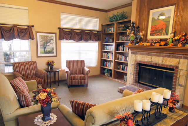 A cozy common living room with sofas and striped armchairs arranged around a stone fireplace and built-in bookshelf, decorated with floral arrangements.