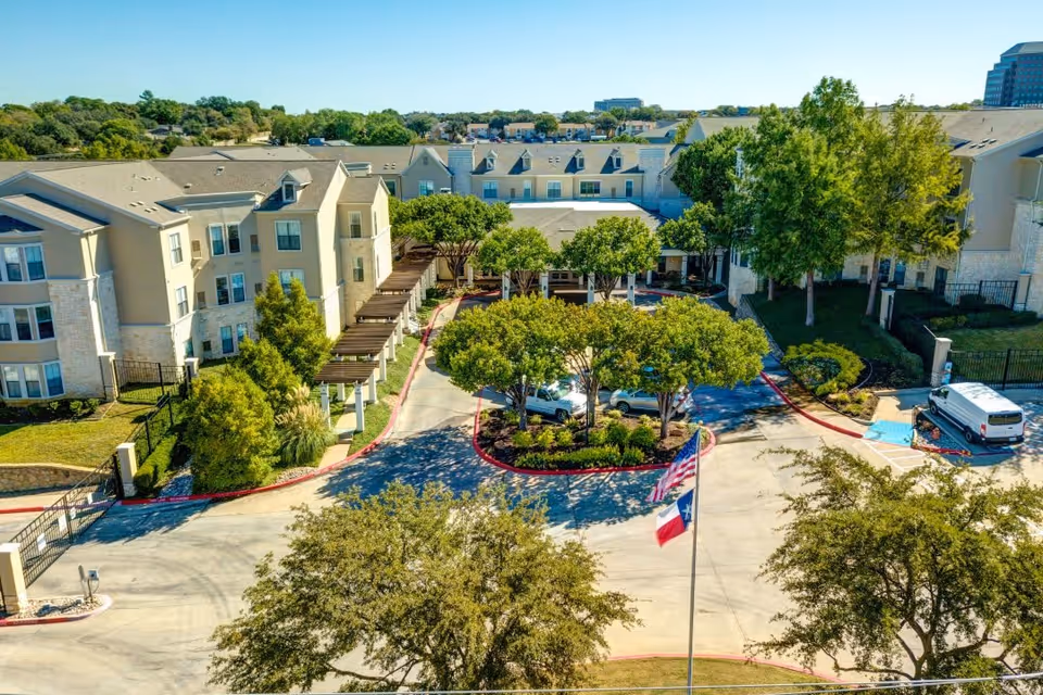 Aerial view of Ridgmar Place senior living facility showing multiple beige buildings with stone accents surrounding a circular driveway with landscaped greenery and trees. Two flags, including the Texas state flag, are visible near the entrance. Several vehicles are parked near the buildings, and the sky is clear and blue.